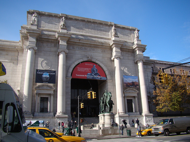 The entrance to the American Museum of Natural History. Clear blue sky, pedestrians walking up the stairs, banners hanging on the facade, and taxicabs in the foreground. Architecture is stately, four tall columns and ornate inscriptions and statues near the roofline.