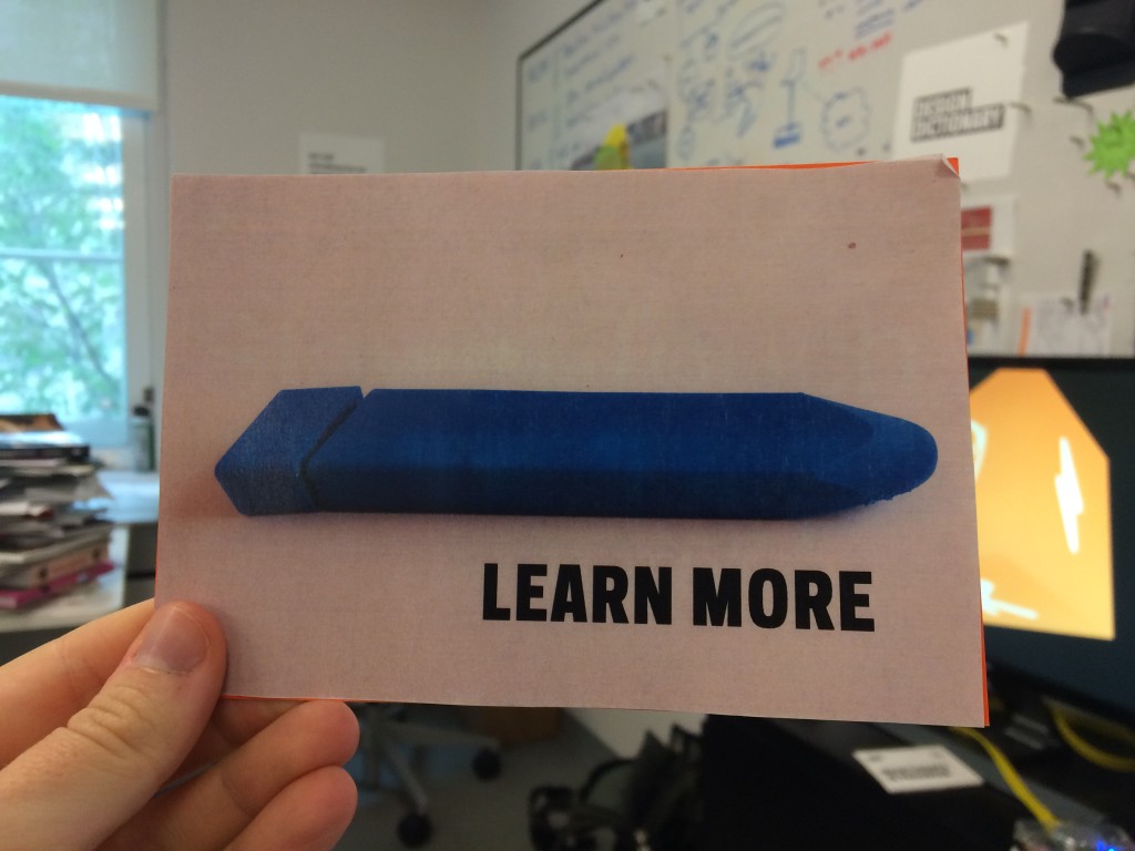 person's hand holding up a postcard in an office setting. postcard says "LEARN MORE" with a blue plastic tube-like object.